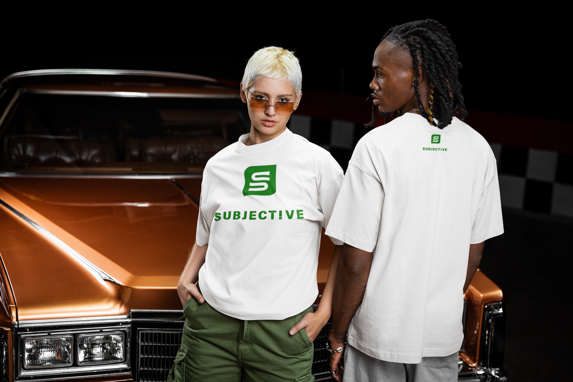 Two people wearing 'SUBJECTIVE' t-shirts standing next to a vintage car.